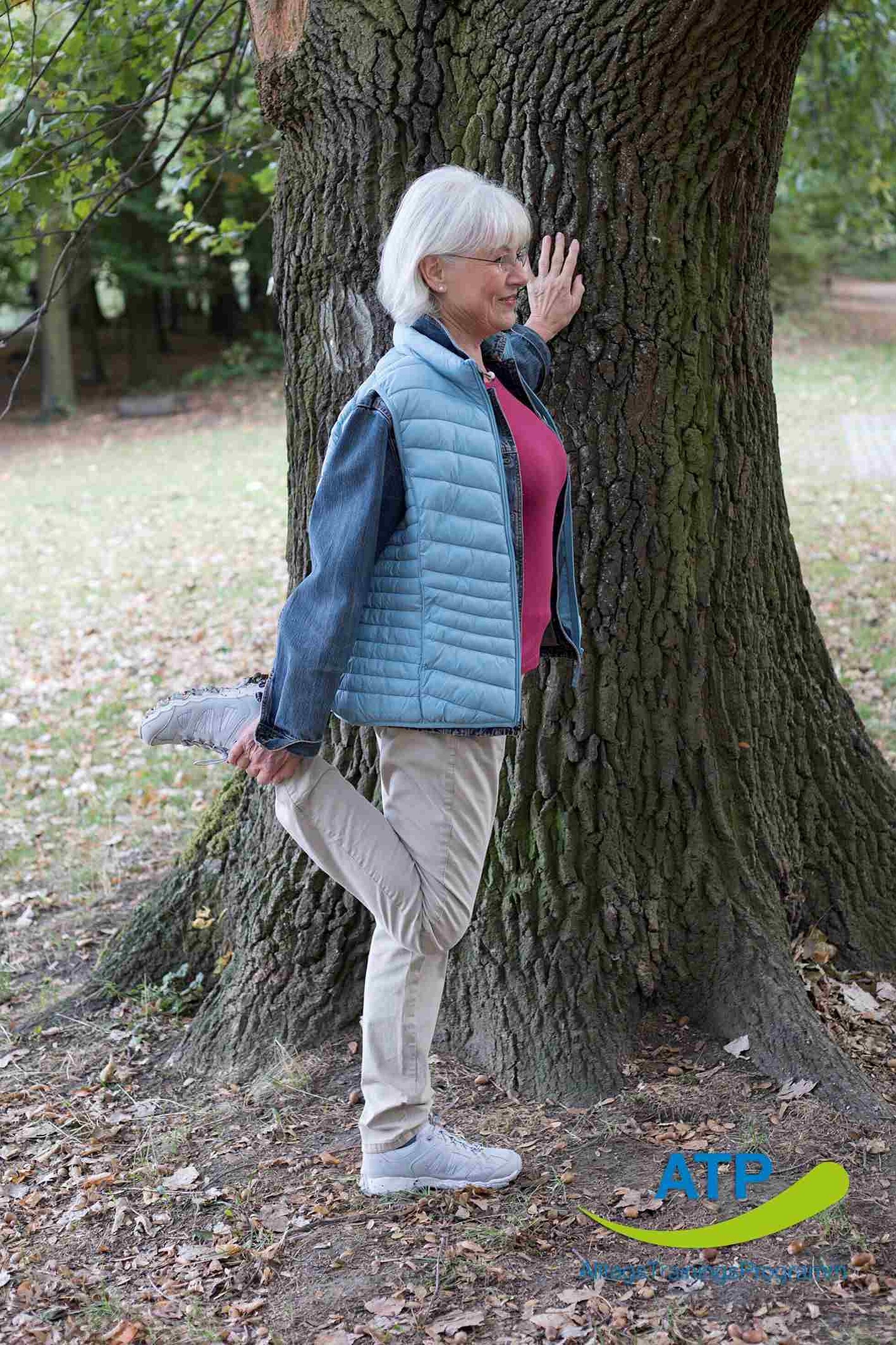 ATP-Dauerangebot Übung 8 Ältere Frau dehnt Oberschenkel und stützt sich an einem Baum ab