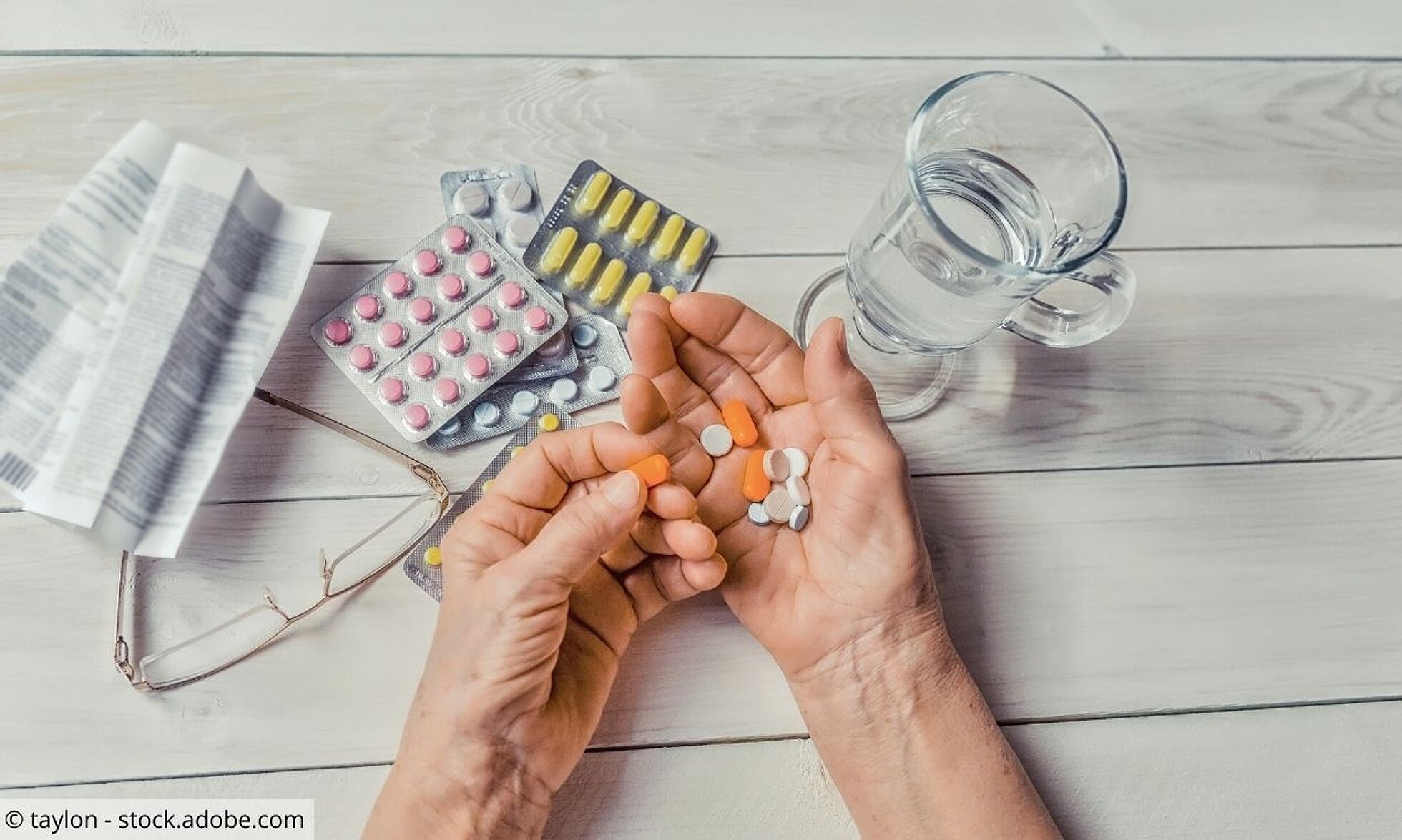 Tisch mit Tabletten, Wasserglas, Lesebrille und Hände einer älteren Person mit Tabletten 