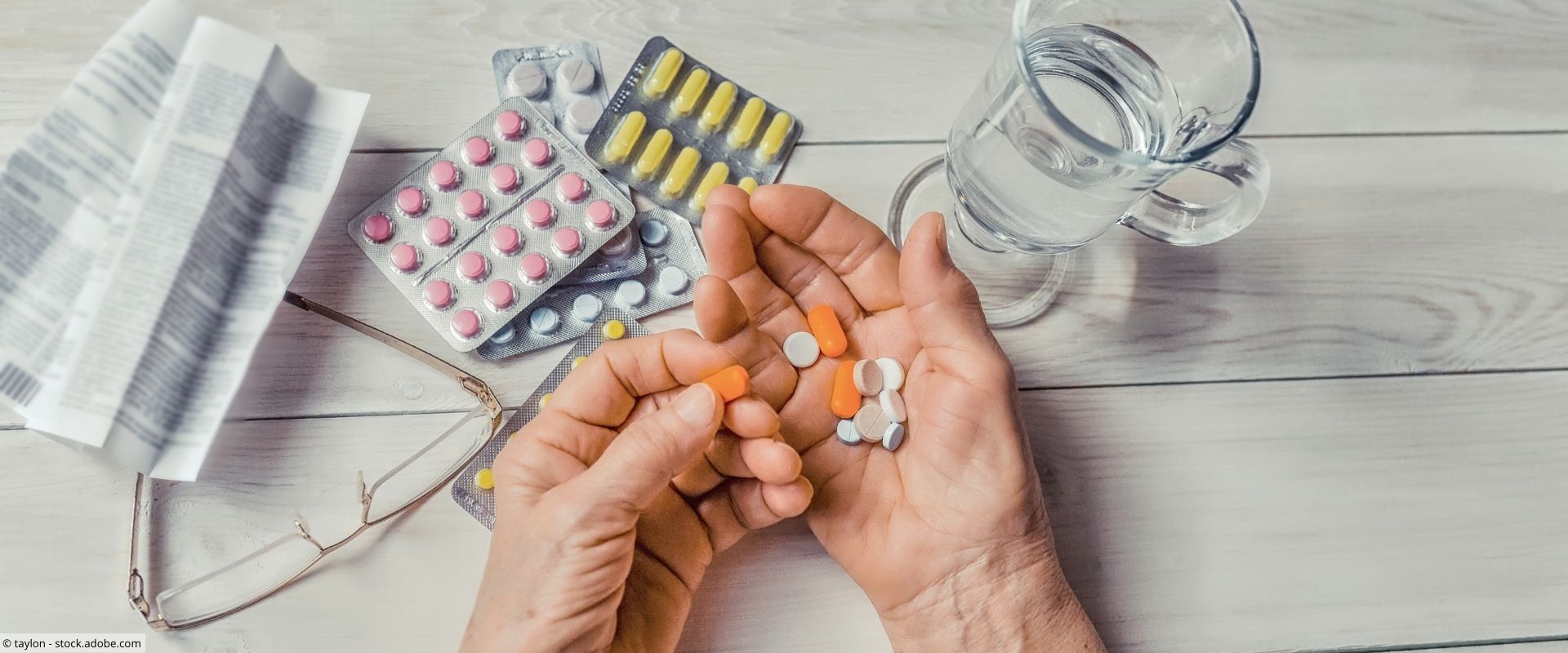 Tisch mit Tabletten, Wasserglas, Lesebrille und Hände einer älteren Person mit Tabletten 